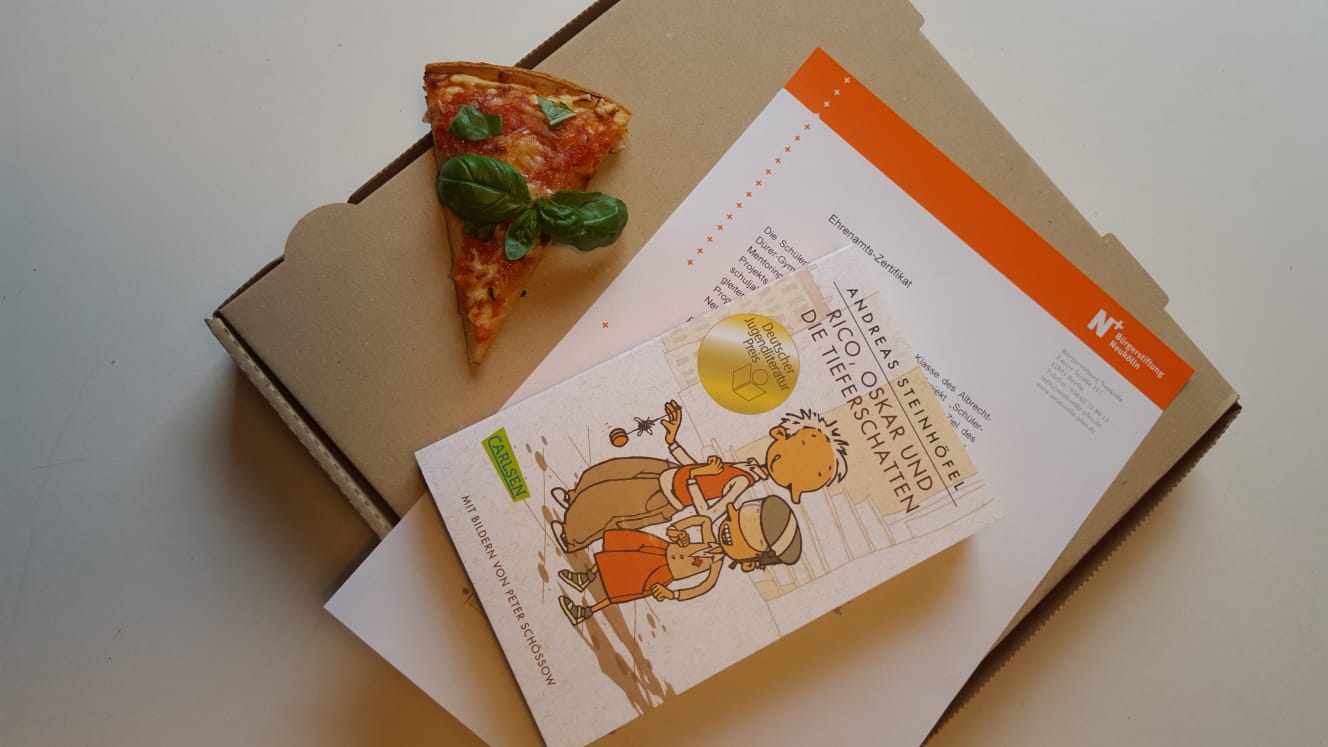 Ein Pizzastück, das Ehrenamts-Zertifikat und ein Buch liegen auf einem Pizzakarton