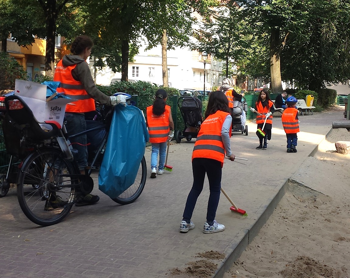 Neuköllner Talente in BSR-Westen räumen mit Besen und Zangen an den Berliner Freiwilligentagen einen Spielplatz auf. Eine Frau mit Fahrrad und blauer Mülltüte begleitet sie.