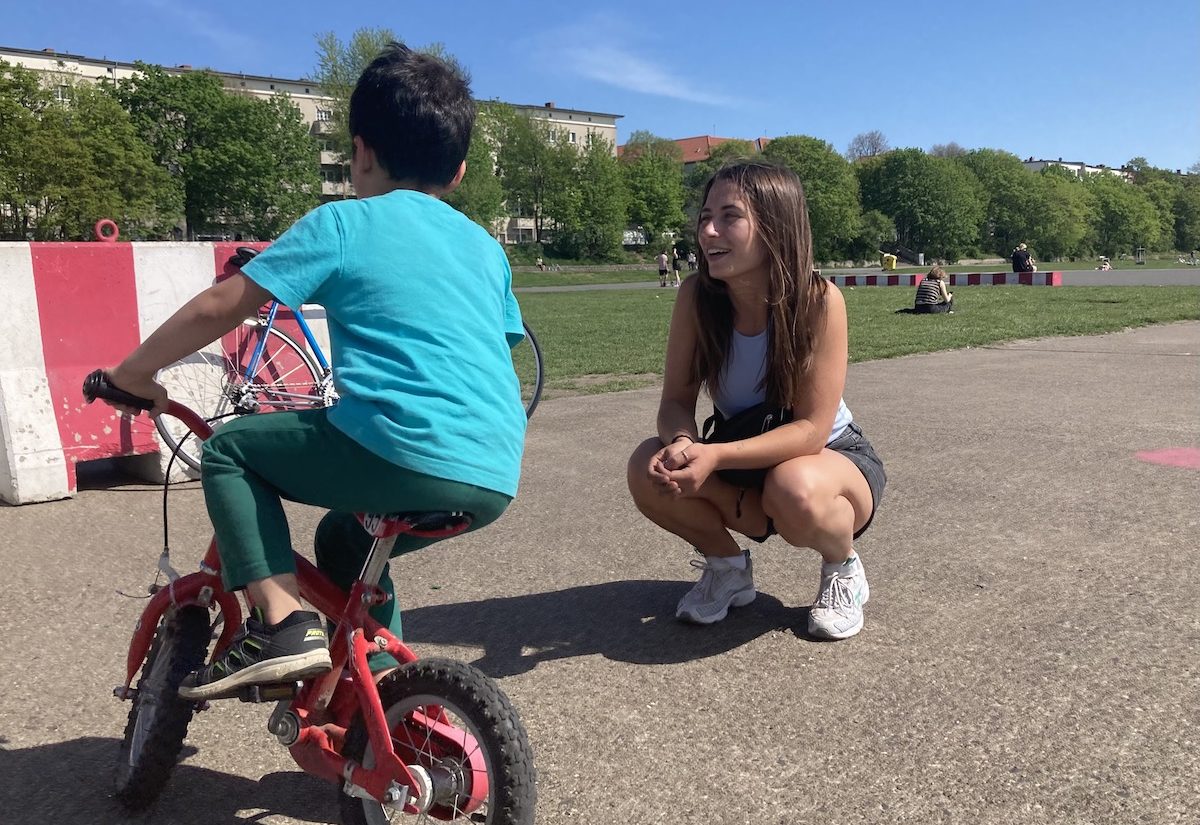 Im Vordergrund Patenkind von hinten auf dem Fahrrad auf dem Tempelhofer, eine junge Frau in der Hocke lächelt das Kind an