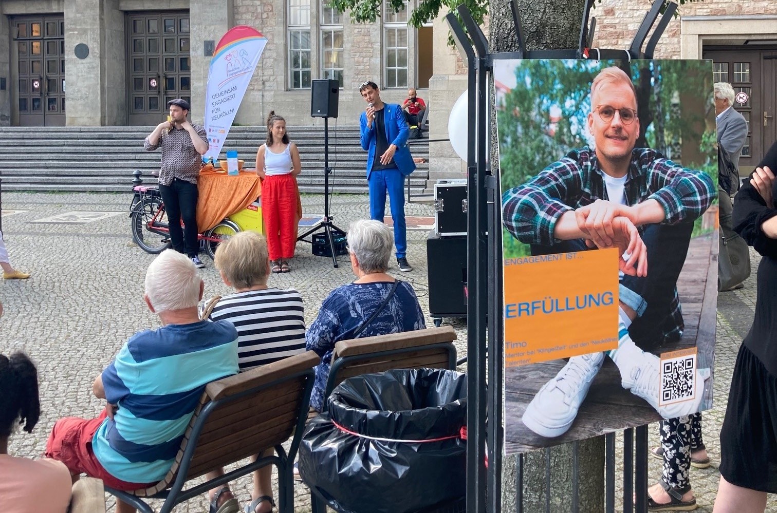 Plakat mit Portrait auf der rechten Bildseite im Vordergrund, im Hintergrund Bezirksbürgermeister Martin Hikel mit Patin im Gespräch