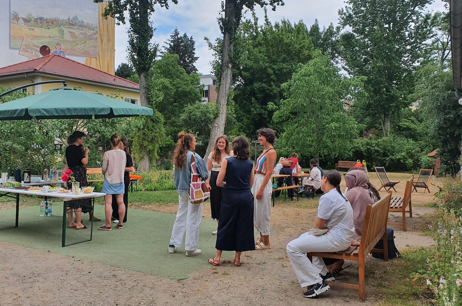 Menschen unterhalten sich in Grüppchen in einem Garten, im Vordergrund links ein Buffet unter einem Sonneschirm