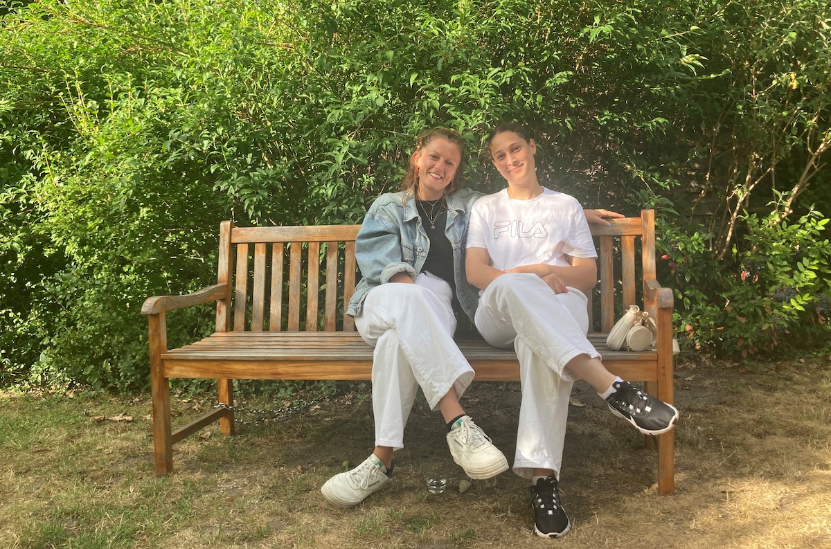 Zwei junge Frauen sitzen auf einer Holzbank vor einem großen Busch, die Köpfe einander zugeneigt