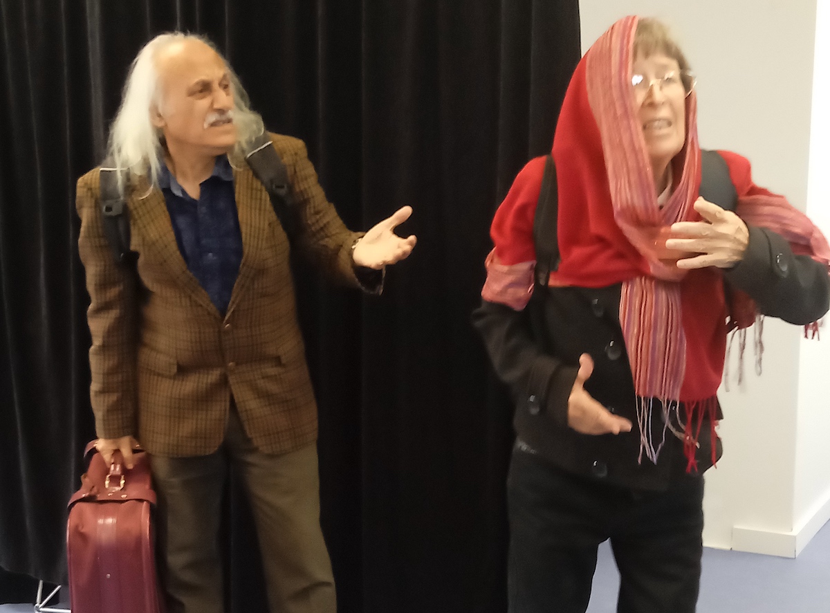 Links steht ein älterer Mann mit Koffer und hebt fragend die linke Hand, rechts vor ihm eine Frau mit Schal über dem Kopf