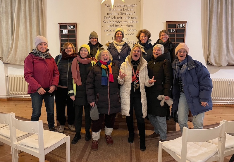 11 Frauen stehen in einer Gruppe zusammen mit den Gesichtern zur Kamera. Einige haben den Mund zum Singen geöffnet.