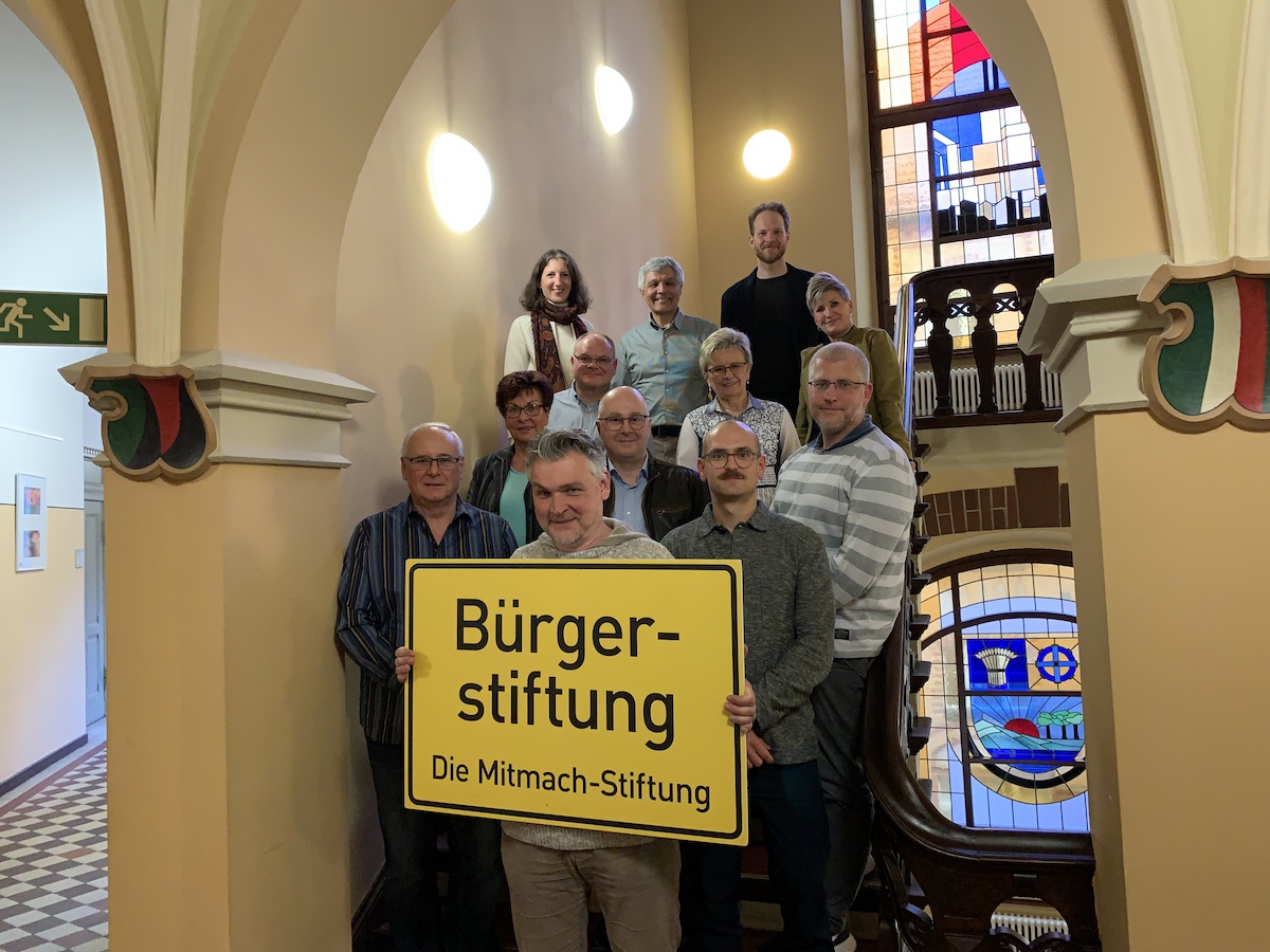 Zwölf Menschen stehen zusammen auf der Treppe im Treppenhaus des Rathaus Lichtenberg. Die vorderen halten ein Schild mit der Aufschrift "Bürgerstiftung. Die Mitmachstiftung."