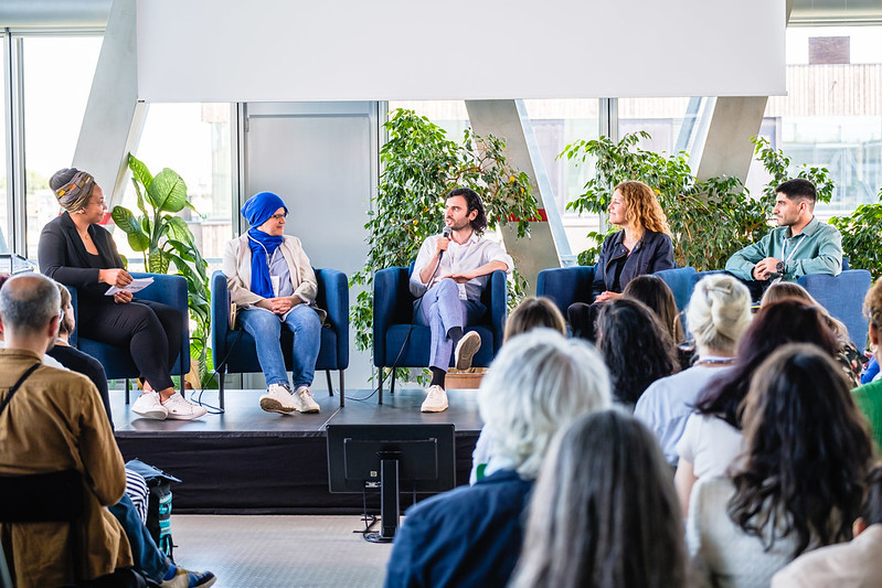 Im Vordergrund sitzen Menschen mit dem Rücken zum Bild. Dahinter sitzen 4 Menschen auf einem Podium mit dem Gesicht zum Bild. Einer hält ein Mikro in der Hand.