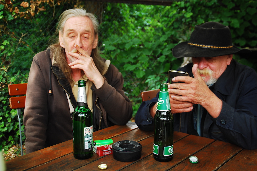 Zwei ältere Männer sitzen im Freien an einem Tisch. Auf dem Tisch 2 Bierflaschen und ein Aschenbecher
