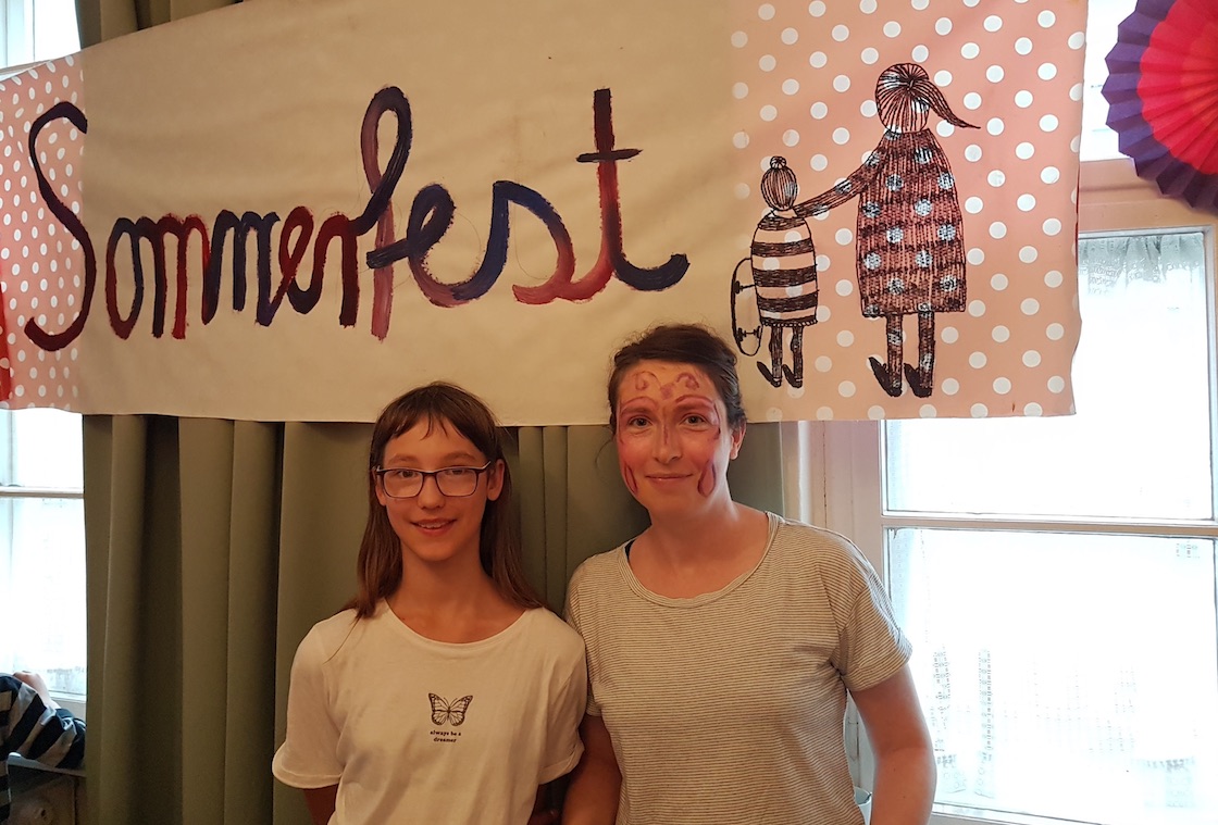 Ein Mädchen und eine junge Frau stehen vor einem Plakat mit der AUfschrift Sommerfest.