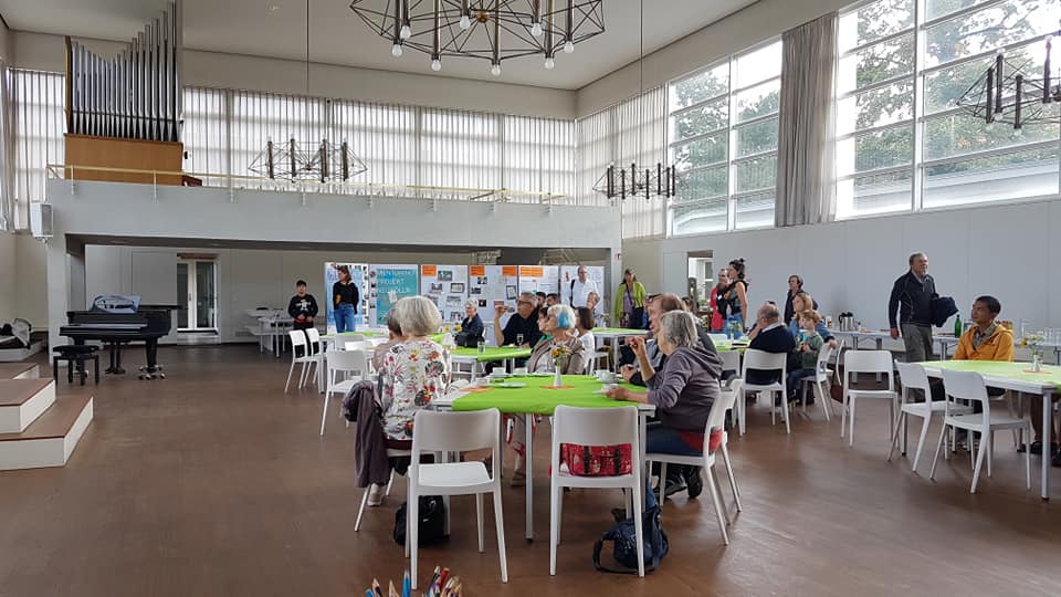 Menschen sitzen in einem Raum an mehreren Tischen, einige stehen vor einer Präsentationswand