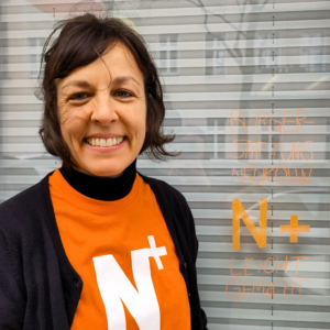 Eine Person mit einem Shirt mit dem Logo der Bürgerstiftung steht lächelnd vor einem Fenster mit dem Logo der Bürgerstiftung darauf.