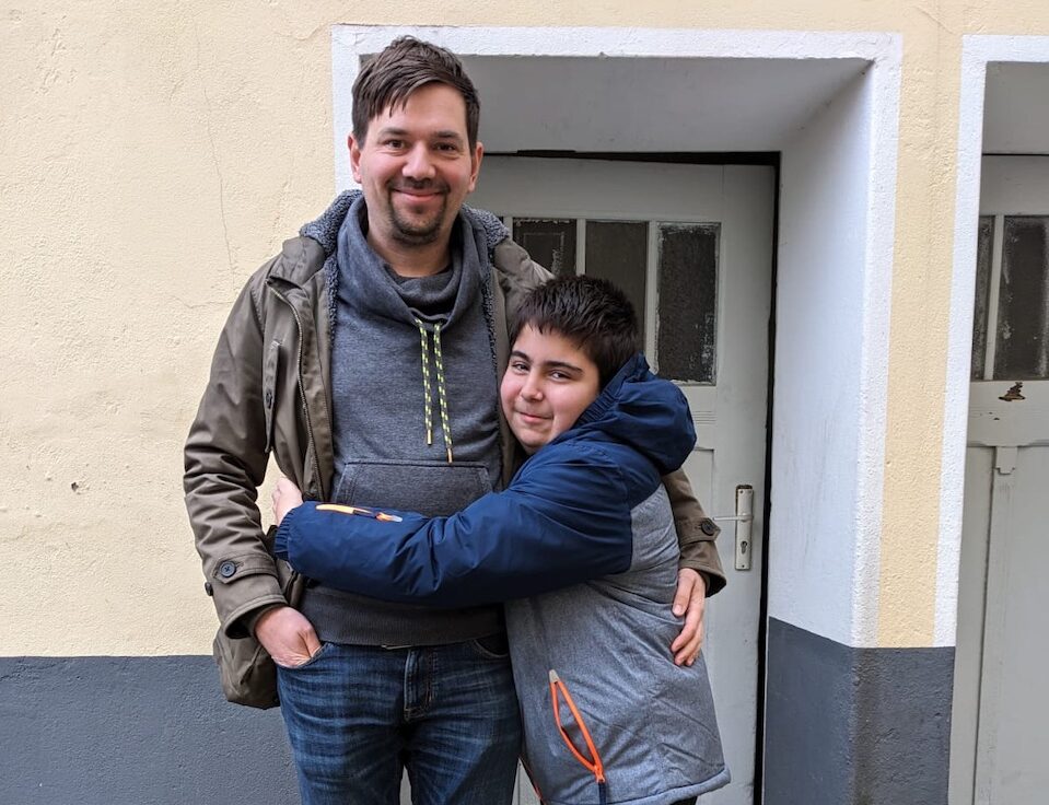 Ein Junge und ein Mann stehen vor einer Haustür. Der Junge umarmt den Mann. Beide lächeln.
