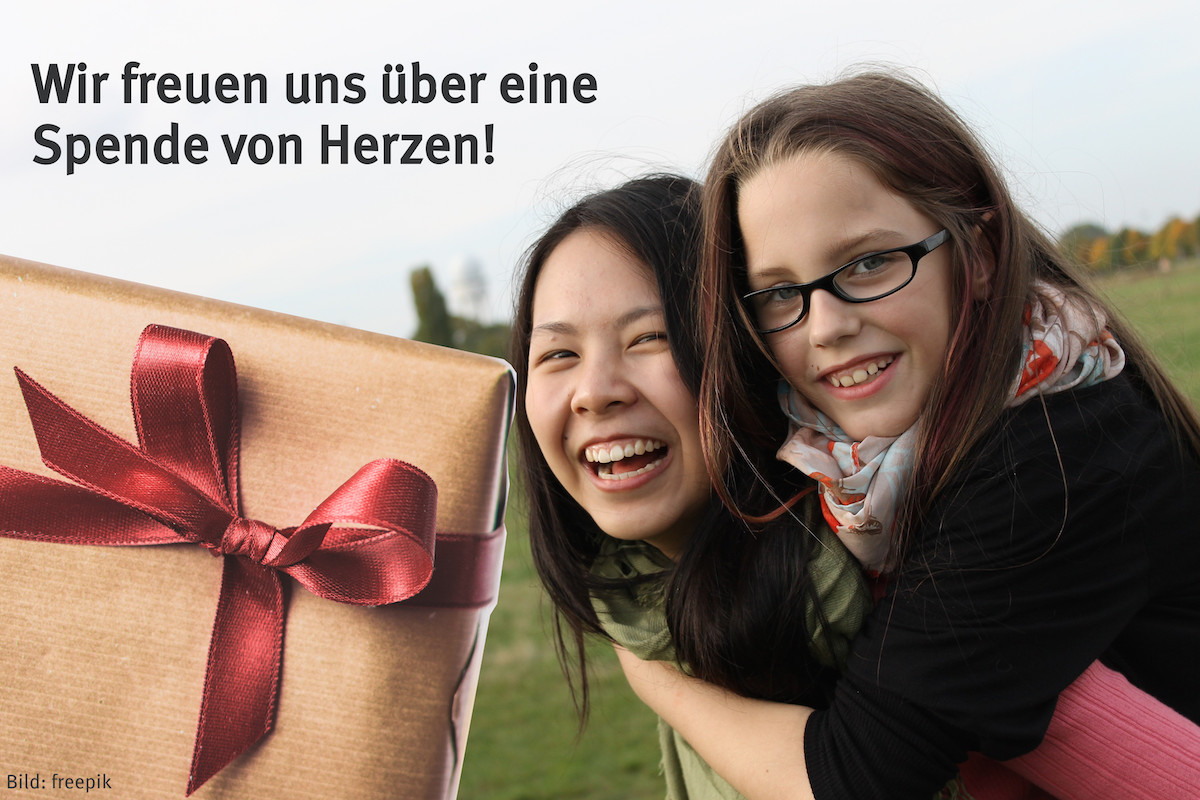 2 Menschen lachen auf der rechten Bildhälfte in die Kamera. Auf der linken Bildseite ist teilweise ein Geschenk mit Schleife zu sehen.