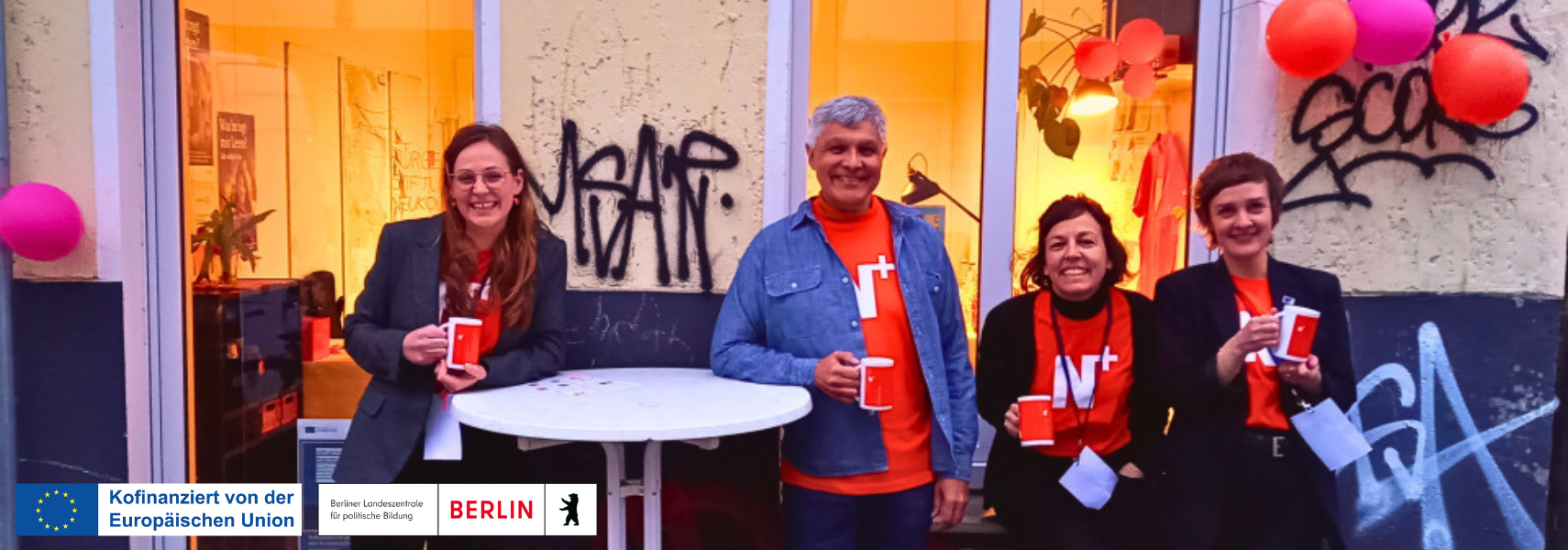 4 Personen stehen vor einem Ladenlokal, es ist eine freundliche Stimmung. Sie tragen T-Shirts mit dem Logo der Bürgerstiftung Neukölln. Logos von EU-Kofinanzierung und Berliner Landeszentrale.